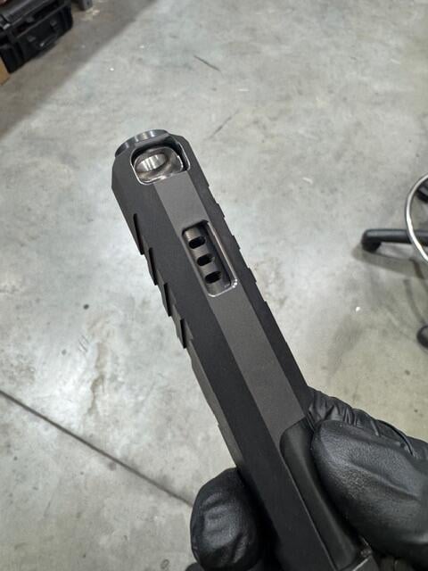 Close-up of a black handgun slide held by a gloved hand, showing a ported window and visible spring inside.